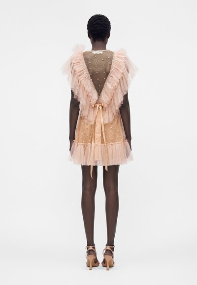 Γυναίκα που φορά διαφανές ροδακινί φόρεμα με διακοσμητικά από πέρλες, μεγάλα πολλαπλά τούλινα βολάν στην πλάτη και δεμένη κορδέλα στη μέση, στέκεται με ψηλοτάκουνα.