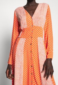 Robe orange ornée de pois blancs et d'un motif floral, avec une patte de boutonnage à l'avant et des manches longues. Le tissu texturé ajoute un intérêt visuel.