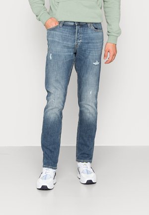 Mężczyzna w wyblakłych, przetartych niebieskich dżinsach, białych tenisówkach i jasnobzielonym zielonym kapturze, stojący na tle jednolitego tła.