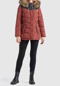 Rød polstret jakke med faux fur hætte, sort bærestykke, lynlås og trykknaplukning, to forlommer, parret med sorte tætsiddende bukser og kampstøvler.