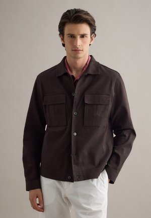 Giovane uomo con indosso una giacca marrone scuro abbottonata sopra una camicia mogano e pantaloni bianchi, in piedi contro uno sfondo chiaro uniforme.