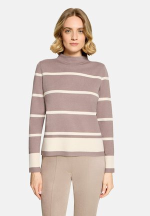 Kvinde med kort blondt hår iført en taupe- og cremefarvet stribet højhalset trøje og beige bukser, stående mod en ensfarvet baggrund.