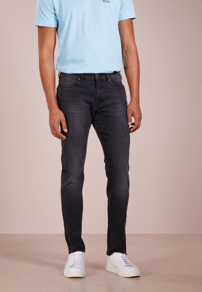 Muž ve slim-fit černých džínách, světle modré košili s krátkým rukávem a bílých teniskách stojí proti jednobodovému pozadí.