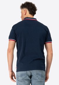 Sötétkék polo ing gallérral, rövid ujjakkal, piros és fehér csíkos részletekkel, és egyenes szegéllyel.