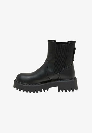 Personne portant des bottines en cuir noir et un vêtement court, debout sur un trottoir urbain ensoleillé près d'une rue et des bâtiments.
