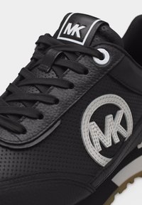 Μαύρο δερμάτινο αθλητικό παπούτσι με τρύπες, μαύρα κορδόνια, λευκό κυκλικό λογότυπο MK στο πλάι και τα αρχικά MK στην ετικέτα της γλώσσας.