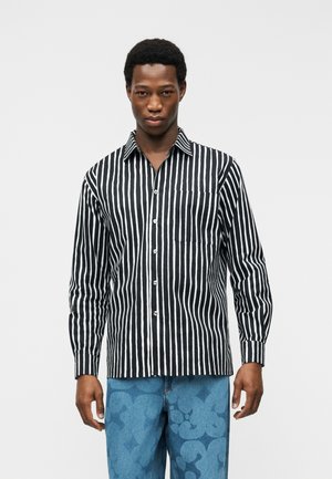 Mężczyzna ubrany w czarno-białą koszulę z pionowymi paskami zapinaną na guziki oraz niebieskie spodnie w wzory, stojący na tle jasnego, jednolitego tła.