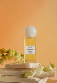 Parfumfles met helder glas, gele vloeistof en een ronde witte dop. Het etiket toont "JAIPUR CHANT" en "SANA JARDIN." Romige bloemige accenten.