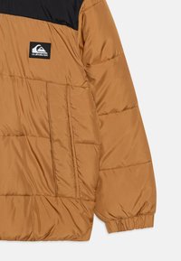 Veste matelassée Quiksilver avec un corps brun et un haut noir. Dispose d'une texture matelassée, de poches latérales et de poignets élastiques.