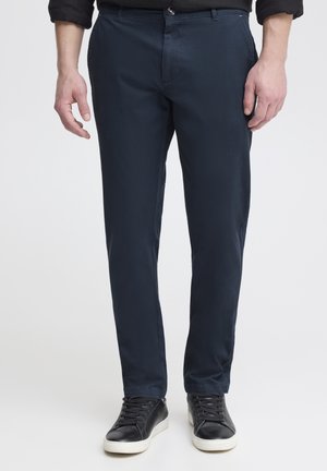 Homme portant un pantalon slim bleu marine foncé et des baskets noires à semelles blanches, debout devant un fond clair uni.