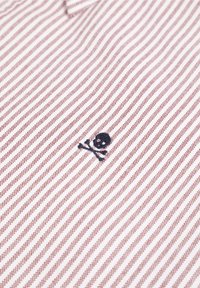 Camisa a rayas con una base blanca y rayas diagonales rojas. Bordado notable de una calavera y tibias cruzadas azules cerca del dobladillo.