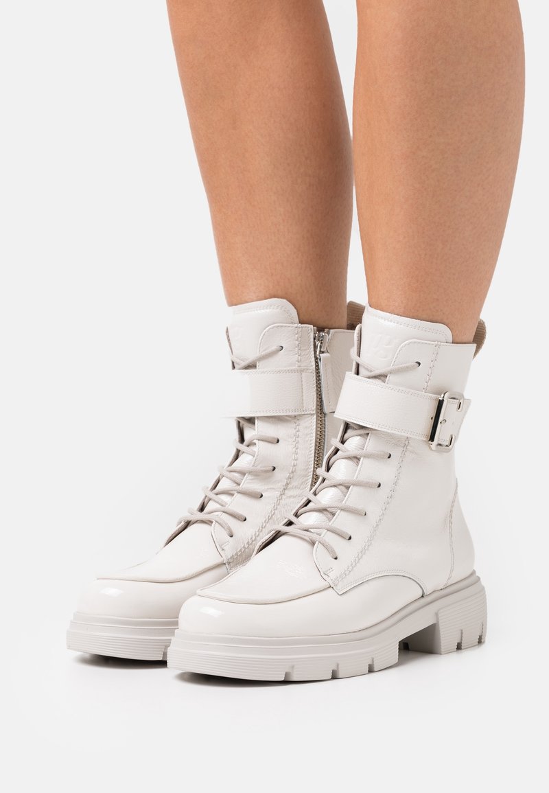 Paire de bottes blanches brillantes à lacets avec semelles épaisses et sangles à la cheville portées sur des jambes nues sur fond blanc.