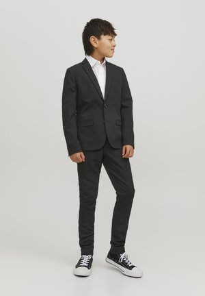 Czarny garnitur z jednokomorową marynarką, biała koszula pod spodem, wąskie spodnie i czarne sneakersy z białymi podeszwami.