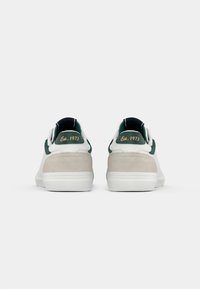 Zapatillas blancas con detalles en verde, talón de ante y texturas contrastantes. Presentan una marca retro "Est. 1973" en la parte trasera.