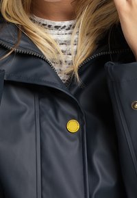 Veste imperméable bleu marine avec une finition brillante, fermeture éclair et un logo de marque circulaire jaune sur la poitrine. L'intérieur présente une couche blanche à motif.