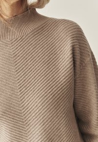 Beige kötött pulóver magas bordázott nyakkal, texturált halszálkás mintával és laza, kényelmes fazonnal.
