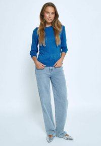 Pulover iz modrega bleščečega materiala s tremi četrtinami rokavov, kombiniran z svetlo modrimi širokimi kavbojkami in srebrnimi balerinkami s konico.