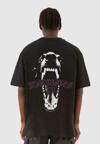 Sort bomulds T-shirt med et stort hvidt grafisk motiv af en hunds åbne mund på bagsiden, med lilla tekst ovenfor grafikken.