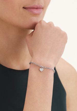 Muñeca de mujer con una pulsera de cadena plateada que tiene un charm en forma de corazón, mano apoyada cerca del mentón, vistiendo una camiseta sin mangas negra.