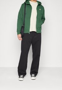 Grön zip-up hoodie med en logo, beige t-shirt, svarta vida byxor och svarta sneakers med vita sålar. Enkelt mönster, avslappnad look.