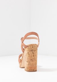 Hnědé semišové sandály na vysokém podpatku s korkovým blokem. Nastavitelný pásek kolem kotníku, otevřená zadní část a zaoblený design špičky. Hladká textura.