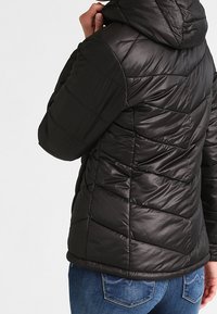 Černá péřová bunda s kapucí, která má prošívaný design s diagonálními švy, hladkou texturu a elastické manžety. Nosit s modrými džíny.