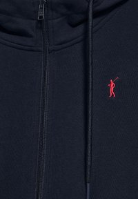 Sudadera con cremallera de color navy hecha de tela suave, con un emblema de golfista rojo en el lado izquierdo del pecho y detalles de capucha con cordones.