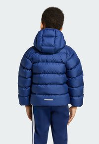 Blå pufferjakke med hette, med horisontale quiltede seksjoner og en reflekterende stripe bak. Glatt, skinnende stofftekstur.