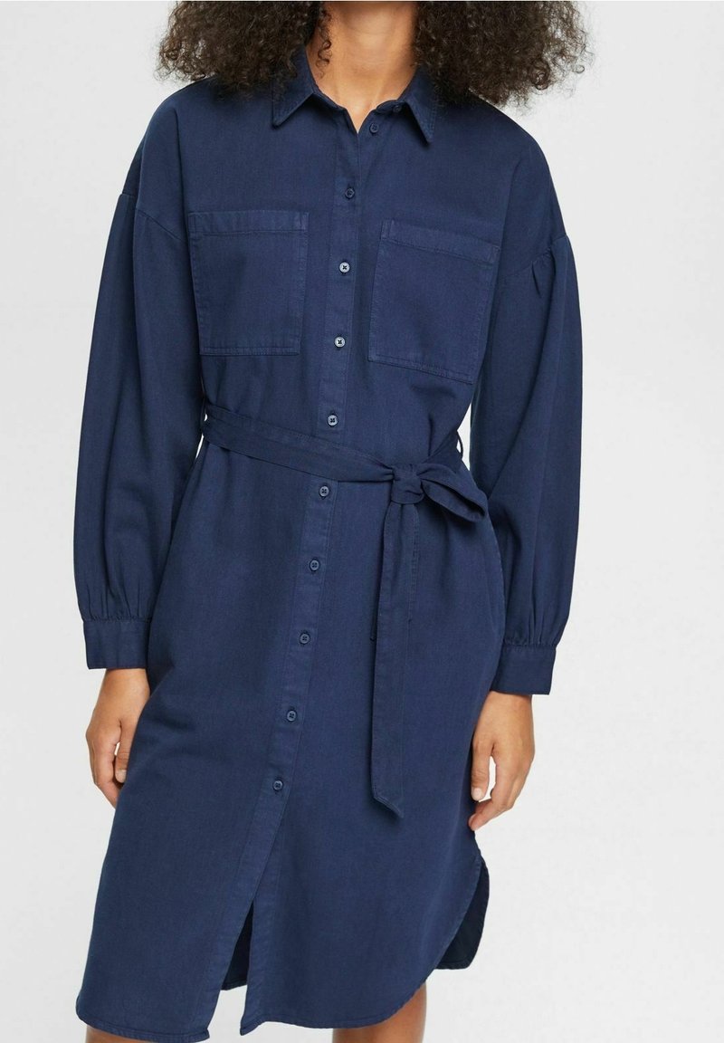 Abito camicia blu navy realizzato in un tessuto morbido, con maniche lunghe, due tasche frontali e una cintura coordinata in vita.