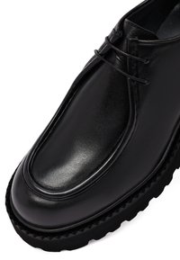 Zapato de cuero negro con un acabado brillante, punta redondeada y una suela gruesa y texturizada. Presenta costuras tonales y cordones en la parte superior.