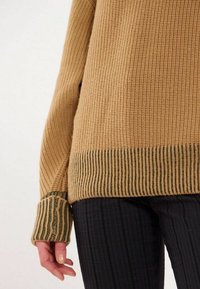 Pull en tricot marron avec une surface texturée, ourlet et poignets rayés verts. Assorti à un pantalon noir à côtes.