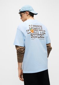 Νεαρό άτομο που φοράει ανοιχτόμπλε καπέλο, γυαλιά ηλίου και στρωμένα μπλε και καφέ μακριά μανίκια με το κείμενο «Always worth the squeeze» και πορτοκαλί γραφικό στην πλάτη.