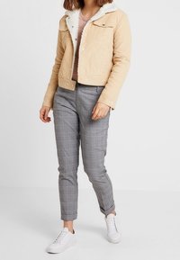 Avskuren beige manchesterjacka med en sherpa-krage, kombinerad med grå rutiga byxor och vita sneakers. Materialet har synlig struktur och sömmar.