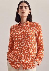 Button-up shirt featuring a white background with large orange polka dots, made of smooth fabric, with long sleeves and a collar.