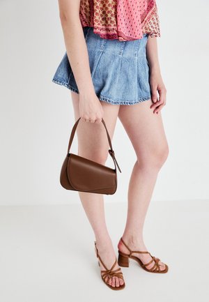 Ruskea keinonahkakäsilaukku, jossa on kaareva muoto ja lyhyt kahva. Laukkua pitää henkilö, joka on pukeutunut farkkushortseihin ja sävyiltään ruskeisiin avokkaisiin.