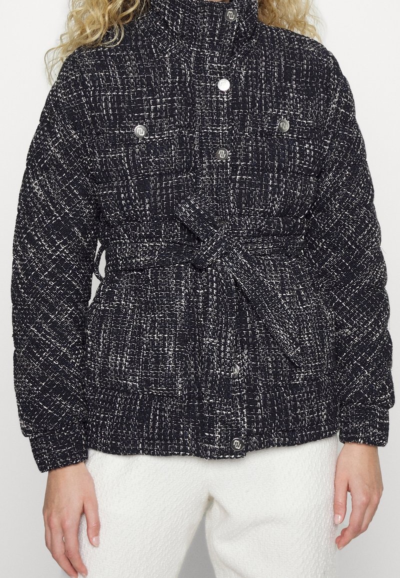 Padded bunda v černobílé tkanině s opaskem v pase, kovovými knoflíky a dvěma náprsními kapsami; má vysoký límec a texturovanou tkaninu.