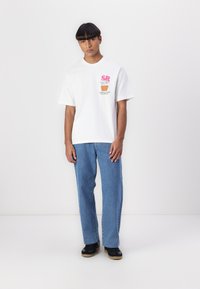 White cotton t-shirt with short sleeves and multicolor graphic print. Paired with light blue denim jeans and black sneakers.