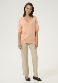Maglione rosa a V con maniche corte, abbinato a pantaloni dritti color beige chiaro e ballerine nere. Texture liscia, design semplice.