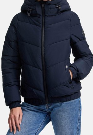 Femme portant une doudoune matelassée bleu marine avec capuche, fermeture éclair devant, poches à boutons-pression et poignets élastiques, associée à un jean bleu.