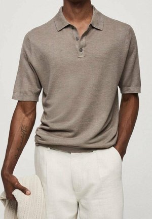 Homme portant un polo texturé taupe à manches courtes rentré dans un pantalon blanc, tenant un pull en laine crème dans la main gauche.