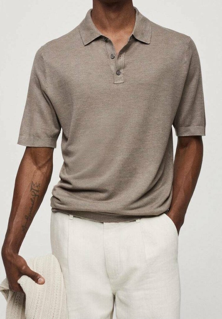 Homme portant un polo texturé taupe à manches courtes rentré dans un pantalon blanc, tenant un pull en laine crème dans la main gauche.