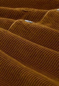 Mustard-colored ribbed fabric with a textured wavy pattern, featuring visible stitching and a subtle sheen from the light.