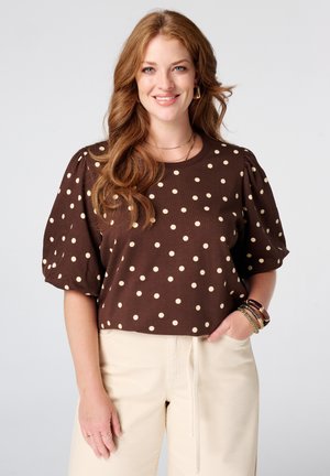 Femme souriante aux longs cheveux ondulés roux portant une chemise marron à pois et un pantalon beige clair avec des bracelets au poignet.