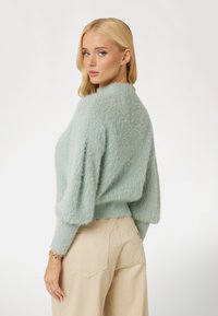 Purūs mėtų žalios spalvos megztinis su aukštu kaklu, laisvu pajūriu ir tekstūruota paviršiumi; derinamas su šviesiai smėliniais aukšto liemens kelnėmis.