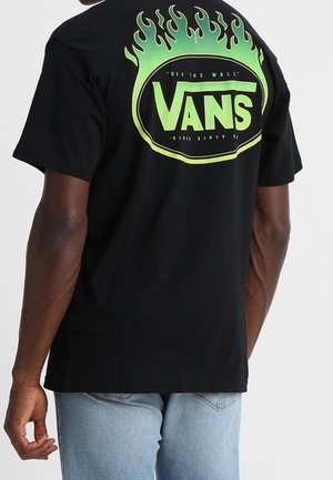 Person iført en sort Vans T-shirt med et grønt flammende logo og teksten "Off The Wall" på bagsiden, kombineret med lyseblå jeans.