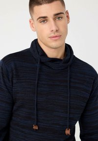 Sudadera con capucha a rayas azul marino y negro con cuello vuelto, tejido texturizado y detalles de cordones. Presenta puntas de cordón de cuero marrón.