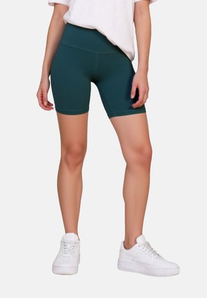 Frau trägt dunkelgrüne Biker-Shorts mit hoher Taille, weiße Sneaker und ein in die Hose gestecktes weißes T-Shirt, steht vor einem einfarbigen Hintergrund.
