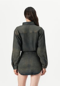 Rochie camasa din denim de culoare verde închis estompat, cu guler, mâneci lungi și un tiv curbat. Detalii cu cusături expuse sunt vizibile.