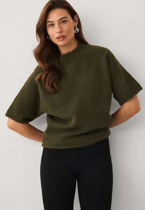 Kvinde med langt brunt hår iført sølvøreringe, en olivengrøn kortærmet sweater og sorte bukser stående mod en lys baggrund.