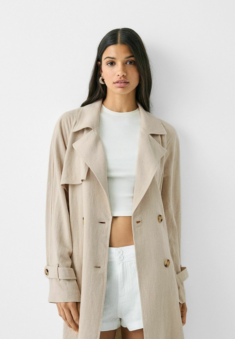 Donna con lunghi capelli scuri indossa un trench beige sopra un crop top bianco e pantaloni corti bianchi a vita alta su uno sfondo bianco uniforme.